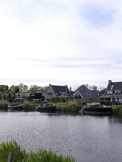 The image depicts a waterway in Oudorp, Netherlands, with modern homes lining the opposite bank. Three boats, all dark-colored, are docked along the bank. Tall reeds and green grasses grow along the water's edge in the foreground. The houses feature dark roofs and varied architectural styles, some with visible windows and chimneys. A Dutch flag is visible between two of the houses. The sky is overcast, suggesting a cloudy day. The water surface is rippled. A wooden post is visible in the extreme lower left corner of the frame.