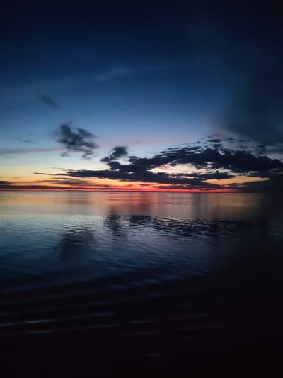 The image depicts a body of water, likely a lake or river, under a twilight sky. The sky transitions from deep blue to a mix of orange and red near the horizon, where the sun has recently set. Dark, puffy clouds are scattered across the upper portion of the sky and clustered along the horizon. The water reflects the colors of the sky and clouds, creating a mirrored effect, with gentle ripples disturbing the reflection. The foreground is obscured in shadow. No people or identifiable objects are visible in the scene. There is no information to confirm that this photo was taken at the city of Mologa, Russia.