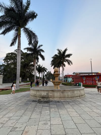 The image depicts a public space in Santa María del Tule, Mexico. The scene includes a stone fountain, palm trees, and a red building labeled "Mercado Artesanias Santa Maria del Tule." People are visible, some sitting near the fountain and others walking on the paved pathway. The sky is a mix of blue and light pink, indicating either sunrise or sunset.