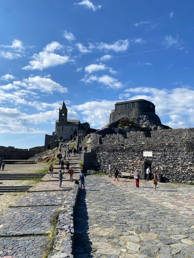 This image depicts a panoramic view of the fortified hillside in Portovenere, Italy, under a bright blue sky with scattered white cumulus clouds. The scene is dominated by historical stone architecture and active pedestrian movement.

In the mid-ground, the Church of San Pietro is prominently positioned on a rocky promontory. It features a distinctive Gothic architectural style with alternating horizontal bands of dark and light stone, and a square bell tower with a conical spire. A wide, ascending stone staircase, paved with irregular dark gray stones, leads towards the church entrance. Approximately 20-30 individuals, casually dressed, are observed walking up and down these steps.

To the right and at a higher elevation than the church, the Doria Castle, a robust, dark gray stone fortress, is visible on a rugged, rocky outcrop. Its thick walls and a rounded section with small windows are evident, indicating its defensive purpose.

The foreground consists of a large, irregularly paved stone plaza or pathway. To the right, a substantial dark gray stone wall, part of the castle complex, features a small arched doorway with a light-colored informational plaque mounted above it. Approximately 10-15 individuals are dispersed across this plaza. One person in a light brown dress walks leftward, while another in red pants stands near a wooden bench. A person in a blue shirt and shorts stands near a low stone retaining wall on the left side of the paved area.

The scene is well-lit by direct sunlight, casting clear shadows, suggesting a mid-day or afternoon time. All major structures are constructed from stone. The environment is an open public space, experiencing moderate foot traffic from visitors.