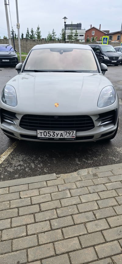 A light-colored Porsche SUV is parked on a paved surface in what appears to be a residential area. The car's license plate reads "T053YA 797 RUS". In the background, there are several houses, trees, and a street lamp. The sky is overcast, suggesting a cloudy day. The overall scene depicts a vehicle parked in an outdoor setting.