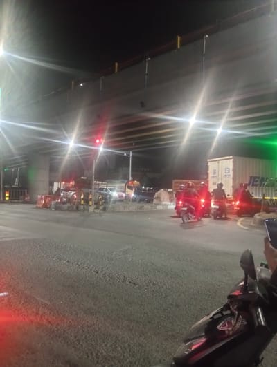 This is a nighttime photograph taken at an intersection in Kedungrejo, Indonesia, likely under an overpass or elevated road. The scene is illuminated by artificial lights, including streetlights and the headlights of vehicles, creating strong lens flares.

The primary subjects are several motorcycles with riders, a truck, and a shipping container. The motorcycles are stopped, some facing away from the camera, with their red taillights illuminated. The riders are wearing helmets. In the background, a large white shipping container with the text "S" and "JEN" is visible, along with other vehicles. Traffic cones and barriers are present, suggesting road work or a controlled intersection.

The time of day is clearly night due to the darkness of the sky and the prevalence of artificial lighting. The weather appears to be clear, as there are no visible signs of rain or fog. The presence of traffic and infrastructure cues like the overpass and intersection suggest an urban or semi-urban environment. The mood is one of waiting, typical of a traffic stop or congestion.

No discernible emotions are conveyed, as the focus is on the objects and the general scene. The visible text includes markings on the shipping container ("S", "JEN") and potentially on the motorcycles, though it's not clearly legible. The overall impression is a busy, illuminated intersection at night, typical of urban Indonesian traffic.