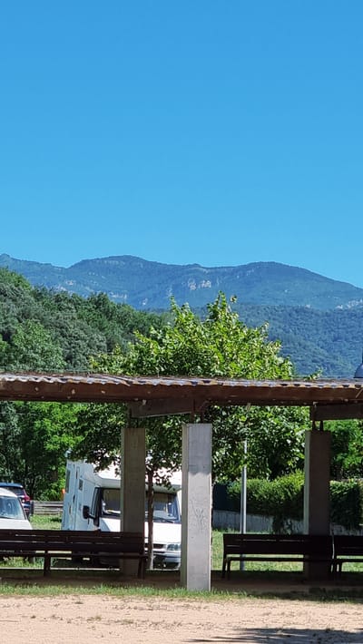 The image shows a clear blue sky above a layered landscape of green trees and distant blue mountains. In the foreground, a wooden structure with a corrugated roof is supported by concrete pillars. Beneath this structure, there are wooden benches and a white camper van. The ground in front of the benches is a sandy or dirt surface, with some grass. The camper van appears to be parked, and its front is visible. The overall scene suggests an outdoor area, possibly a rest stop or campground, with a natural backdrop of hills and mountains.