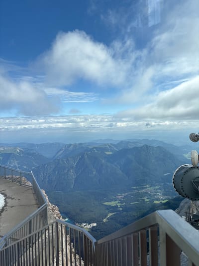 The image depicts a panoramic view from a high vantage point overlooking a mountainous landscape and a village. In the foreground, a metal railing and a concrete walkway with a small patch of snow indicate a man-made structure designed for observation. The walkway curves to the left, suggesting a pathway or viewing platform.

The midground reveals a vast expanse of layered mountains, with the closest peaks covered in dark green forests and receding into hazy blue hues in the distance. A body of water, appearing as a turquoise lake, is visible nestled within the valley floor, surrounded by dense forest. A small cluster of buildings, suggesting a village or settlement, is situated near the lake.

In the upper right portion of the image, large, industrial-looking satellite dishes or communication equipment are prominent. These objects are gray and round, with visible circular detailing.

The sky above is a bright blue, dotted with scattered white clouds, some of which are wispy and others more dense and voluminous. The overall impression is one of a clear day with excellent visibility, offering a grand vista of the natural environment and a human settlement. The scene appears to be from a mountainous tourist destination, possibly a cable car station or a summit observatory.

While the image provides a clear view of the landscape, the specific location in the city of Ehrwald, Austria, cannot be definitively confirmed from the visual information alone. However, the mountainous terrain, lake, and village are consistent with the geography of the Austrian Alps. There are no people visible in the image.