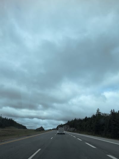 This image shows a view from a vehicle on a highway in Lepreau Parish, Canada, under a cloudy sky. The highway is paved with asphalt and has white lane markings. On the left side of the road, there is a grassy embankment with some trees and rocks. On the right side of the road, there is a dense forest. A silver trailer is attached to a vehicle in the distance, traveling in the same direction. The overall atmosphere is overcast and somber.