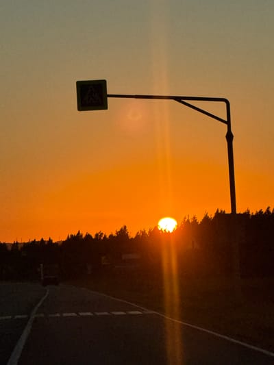 The image depicts a sunset scene on a road in Morozovka, Russia. The foreground features a dark asphalt road with white markings, suggesting it's a highway or main road. To the left, a white line delineates the edge of the road. In the mid-ground, a silhouette of trees forms a dark line against the vibrant orange and yellow sky. The sun, a bright, glowing orb, is partially obscured by the trees on the right side of the image, casting a strong beam of light vertically upwards. A pedestrian crossing sign on a pole is silhouetted against the sky, positioned to the left of the sun. The sky transitions from a light yellow at the horizon to a pale blue-grey at the top. A single vehicle is faintly visible on the road in the distance to the left. The overall impression is one of a rural or roadside setting during the golden hour.