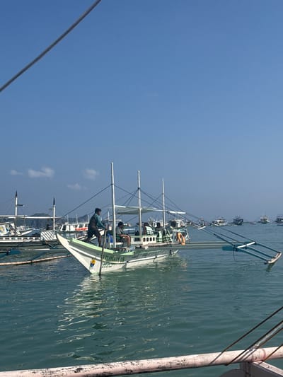 The image shows a fleet of small, traditional Filipino outrigger boats, known as 'bangka,' moored in the waters of El Nido, Philippines. A man is standing on one of the boats, using an oar to maneuver it. Additional people are seated on the boats in the distance. The water is calm, reflecting the clear blue sky above. In the foreground, a portion of a weathered wooden post is visible.