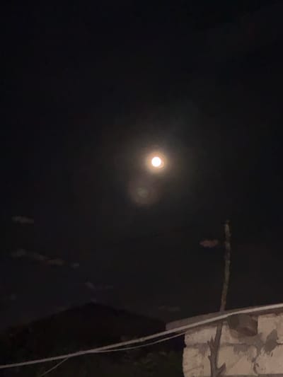 This image captures a nighttime scene dominated by a dark sky, with a bright, luminous full moon at its center. The moon is the primary subject, emitting a strong glow that creates a lens flare effect, appearing as a bright white orb with a softer halo. To the right of the moon, a fainter, slightly blurred circular light is also visible, possibly another celestial body or a reflection.

The foreground is mostly obscured by darkness, but hints of elements suggest an outdoor setting. On the far right, a vertical structure appears, possibly a building or a utility pole, made of rough, textured material, possibly stone or concrete, rendered in muted beige and grey tones. A thin, white cord or cable hangs vertically alongside this structure. In the lower portion of the image, some darker shapes and textures can be discerned, suggesting foliage or debris on the ground, but these are indistinct. A horizontal, dark, branch-like object stretches across the mid-ground of the image, silhouetted against the dark sky.

The overall impression is one of a clear night, with the moon being the dominant source of light. The setting appears to be somewhat rural or suburban, given the presence of the rough-hewn structure and the natural elements. The time of day is clearly night, and the scene evokes a sense of stillness and quiet. There is no visible text in the image.