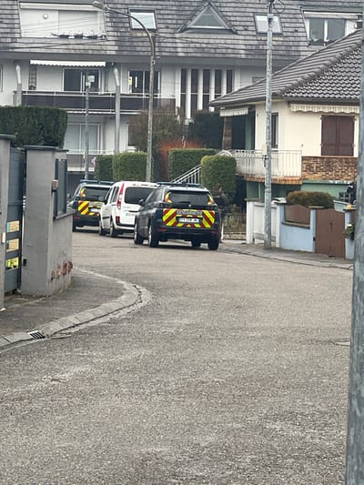 The scene depicts a residential street in Ittenheim, France, with three vehicles parked consecutively on the right side of the asphalt road, facing away from the viewer. The rearmost vehicle is a dark Gendarmerie SUV, displaying yellow "GENDARMERIE" text and red/yellow chevron markings, with license plate FY 528 JN. Immediately in front of it is a white van, showing a partially visible license plate beginning with DZ-61..., followed by a second dark Gendarmerie SUV.
A person in dark clothing, consistent with a Gendarme uniform, is visible standing near the front Gendarmerie vehicle, partially obscured by vegetation and a residential structure on the right side of the street.
The environment includes residential buildings lining the street. On the left, a multi-story house features a dark shingle roof, white facade, multiple windows, and balconies, with one upper window showing a rolled-up exterior blind. On the right, a house with a tiled roof, cream-colored exterior, and brown shutters stands behind a white-railed balcony; its lower facade is green. Trimmed hedges border the properties. A prominent streetlamp with a curved arm is centrally positioned in the mid-ground. The paved street includes a curb and a storm drain on the left side. A grey metal gate is partially open on the left foreground, and a brown wooden gate is visible on the right. The sky is overcast.