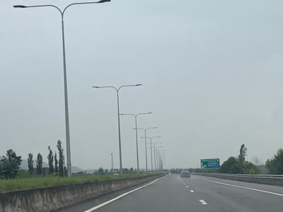 The image depicts a highway scene in the vicinity of ნიგოითის თემი, Georgia, under an overcast sky. A line of streetlights extends along the right side of the road. A concrete barrier separates the highway from a grassy embankment with a row of tall, slender trees. In the distance, a green directional sign is visible, indicating "BATUMI" and "UGUTI" with arrows. A dark-colored car is driving on the highway ahead, and the road appears wet, suggesting recent rain. The overall atmosphere is muted due to the cloudy weather.