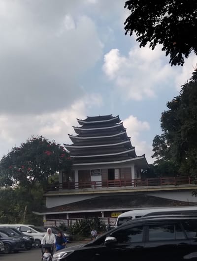 In the city of Gudangkahuripan, Indonesia, a multi-tiered pagoda-style building with dark, curved roofs and white trim stands prominently. Trees with red flowers and lush green foliage surround the base of the building, with a dark, leafy tree canopy overhead. In the foreground, several parked cars are visible, including a black SUV in the immediate foreground. Two women, one wearing a white hijab and the other a blue outfit, are walking in front of a stroller containing a child. The sky is overcast with patches of blue peeking through.