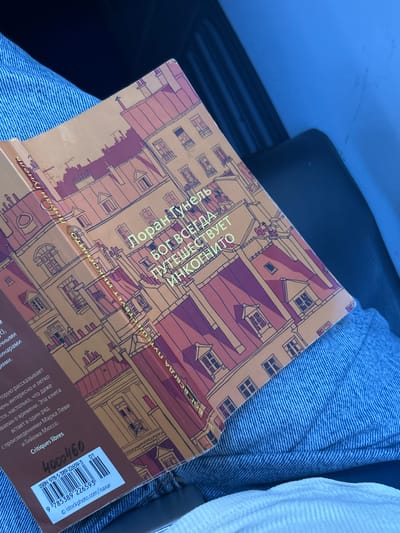 The image shows a close-up of a book titled "БОГ ВСЕГДА ПУТЕШЕСТВУЕТ Инкогнито" (God Always Travels Incognito) by Лоран Гунель (Laurent Gounelle). The book's cover features an illustration of a European-style building with many windows. The book is lying on top of blue denim fabric, likely jeans, and a white ribbed fabric, possibly a shirt or blanket.
The book is positioned diagonally across the frame, with its spine facing upwards. A barcode sticker with the ISBN 978-5-389-22659-3 and "Critiques libres" is visible on the bottom right corner of the book's back cover. Some text in Russian is also visible on the book's cover, describing its content as an interesting and easy read that stands alongside works by popular authors like Mark Levy and Guillaume Musso.
The lighting suggests it might be daytime, possibly indoors or in a shaded outdoor area, given the soft illumination and lack of harsh shadows. The overall impression is casual, perhaps indicating the book is being read or transported by someone in everyday clothing. The focus is clearly on the book and its title, suggesting it's the primary subject of interest. There are no people prominently visible, but the context implies human presence through the clothing and the book itself.