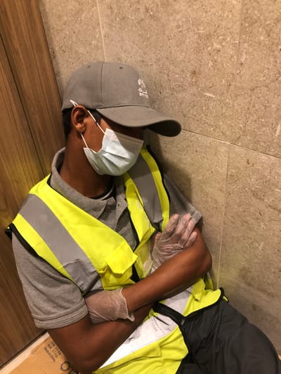 A person wearing a grey cap, a light blue face mask, a grey shirt, a yellow reflective safety vest, and clear plastic gloves lies on a floor with their arms crossed. The floor is partially tiled with a light beige stone pattern and partially made of light brown planks. A portion of a cardboard box with printed text is visible near the person's side. The person appears to be resting or sleeping.