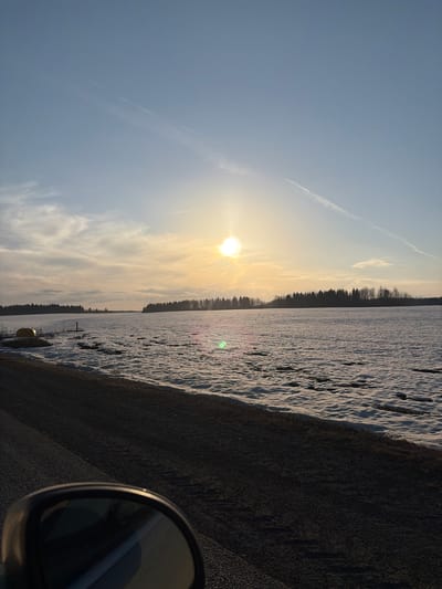 The image shows a winter landscape with a snow-covered field under a sunny sky. A dark line of trees is visible on the horizon, separating the field from the sky. The sun is shining brightly, causing a lens flare. A road is visible in the lower part of the image, covered in dark material. A side mirror of a vehicle is present in the bottom right corner. The location is Bol'shaya Sosnova, Russia.