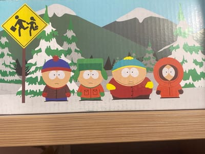 The image shows a detailed illustration of four South Park characters: Stan, Kyle, Cartman, and Kenny, standing in front of a snowy mountain landscape. A yellow diamond-shaped sign with the silhouette of two children walking with briefcases is prominently displayed on the left. The characters are depicted in their typical attire. The illustration appears to be printed on a box or a similar item, with a wooden surface visible at the bottom of the frame. There is no discernible connection to the city of Obninsk, Russia, or any urban environment.