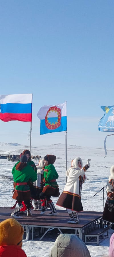 The image depicts an outdoor cultural event occurring on a snow-covered, flat expanse under a clear blue sky in Nelmin-Nos, Russia. In the mid-ground, a group of approximately five to six individuals, dressed in traditional indigenous attire featuring green tunics, brown skirts/trousers, and fur-trimmed hoods/hats, are performing on a dark-colored elevated stage. One female performer in a white fur-trimmed coat and brown skirt is prominently featured on the right side of the stage, with her right arm extended upwards. Behind the stage, the Russian national flag (white, blue, red horizontal stripes) and the flag of the Nenets Autonomous Okrug (white and light blue with a circular emblem) are displayed on poles, alongside a partially visible third flag depicting a white reindeer and fish on a light blue background. In the background, a herd of reindeer is visible against the vast, snowy landscape, which includes distant low hills. The foreground shows the top of the heads and shoulders of at least two spectators, one in an orange hat and red jacket, and another in a grey hooded jacket, indicating an audience is present. A microphone stand is partially visible on the right side of the stage. The scene is brightly lit by natural light.
