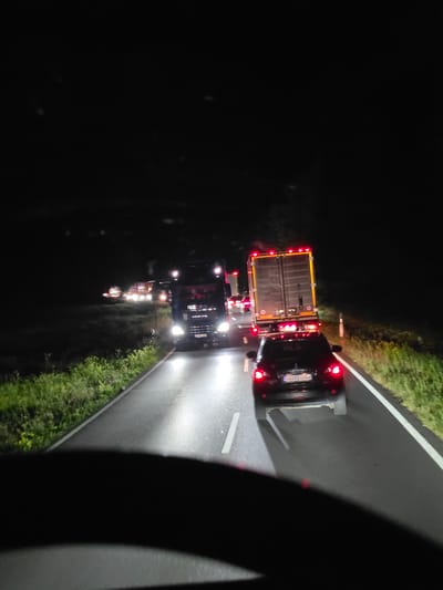 The image depicts a nighttime traffic scene on a road in or near Seligenstadt, Germany. Multiple vehicles are visible, including trucks and cars, driving in the same direction on a paved road. The road is marked with white lines. Green grass and vegetation are present on the sides of the road, suggesting a rural or suburban environment. The vehicles have their headlights and taillights illuminated, indicating low visibility. The overall impression is of a slow-moving or stopped traffic situation. No people are clearly identifiable.