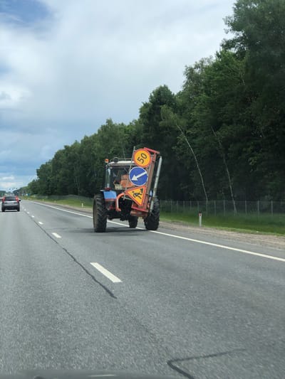 A blue tractor with construction signs is driving on a highway. One sign indicates a speed limit of 50 kilometers per hour. Another sign shows a blue arrow pointing to the right, and a third sign depicts a person working. The tractor is carrying a red trailer attached to its rear. The tractor has a license plate with the number "8091 CA 67". The scene takes place on a cloudy day on a paved highway. Trees line the side of the road, and a wire fence separates the road from the trees. A silver car is visible driving ahead of the tractor. The location is Kablukovo, Russia.