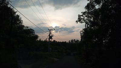 The image depicts a rural road in Keudesimpangempat, Indonesia, during twilight. The sun is low in the sky, partially obscured by clouds, casting a soft glow through a gap. Overhead, power lines crisscross the frame, supported by a utility pole with a transformer. Dense vegetation lines both sides of the road, with a mix of trees and smaller plants. In the distance, a dark-colored truck is visible on the road, and a motorcycle with its headlight on is passing it. The overall atmosphere is serene and dusky, characteristic of the end of the day in a village setting.
