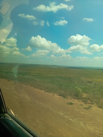 The image captures a view through a window, likely from a vehicle, looking out onto a vast, flat, grassy landscape under a bright blue sky dotted with white, puffy clouds. In the foreground, a dirt road or path runs diagonally across the frame, with some blurred green vegetation visible on its edges. The middle ground consists of rolling hills covered in green and yellowed grass, indicating a rural or steppe environment. In the distance, a faint line of trees and some small structures, possibly buildings, are visible on the horizon. The scene is characterized by open space, natural elements, and a sense of distance. The location is identified as Aleksandrovka, Kazakhstan.