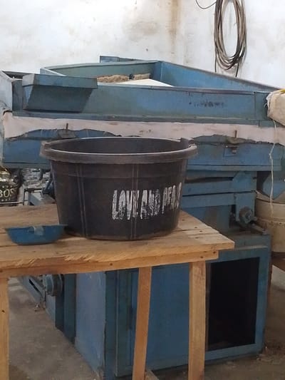 The scene depicts an industrial indoor environment in Olomoda, Nigeria, featuring a large, blue processing machine dominating the background. This multi-level machine has chutes and appears to handle a fibrous, light brown material on its upper surface. A light-colored fabric or tarp is draped across the front of the machine. In the midground, a simple wooden table with visible plank construction holds a large, dark plastic bucket bearing the white inscription "LOVE AND PEACE" on its side. To the left of the bucket on the table, a small, light blue, shallow bowl is present. The background wall is light-colored with some discoloration, and coiled cables or hoses hang from it in the upper right. A partial view of a bag with "BOS" text is visible behind the table to the left. No individuals are present, and no actions or interactions are depicted. The floor is a concrete surface.