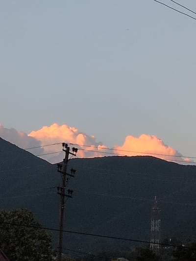 The image shows a landscape scene, likely in Şəki, Azerbaijan, during sunset. The sky is a pale blue, with a line of clouds illuminated by the setting sun, casting a warm orange and pink glow. These clouds are positioned along the silhouette of a dark, mountainous terrain that dominates the right side of the frame. A prominent feature in the foreground is a utility pole with wires, suggesting an inhabited or semi-rural area. The pole is dark and stark against the lighter sky and the shadowed mountain. In the distance, on the mountain slope, a tall, lattice tower is visible, possibly a communication tower or pylon, hinting at infrastructure in the area. The overall mood is serene and picturesque, typical of a twilight landscape. There is no visible text in the image.