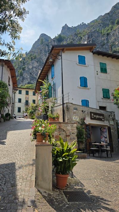 The image depicts a narrow, cobblestone street in Limone sul Garda, Italy, lined with traditional buildings and leading towards a steep, rocky mountainside. In the foreground, a large terracotta pot overflows with vibrant pink and white flowers, placed on a stone and stucco base. To the right, a building with a white facade features blue shutters and a sign reading "L'Angolo Rustico Bar". A small outdoor seating area with a table and two chairs is visible in front of the bar. Further up the street, other buildings with light-colored facades and green or blue shutters are visible. Two individuals are walking up the street, and another person is seated on a low wall on the left side. Lush greenery and flowering plants adorn the buildings and pathways, enhancing the picturesque Mediterranean atmosphere. The sky is clear with scattered clouds, and the sun casts long shadows across the cobblestones. The mountain backdrop dominates the upper portion of the image, showcasing its rugged texture and sparse vegetation.