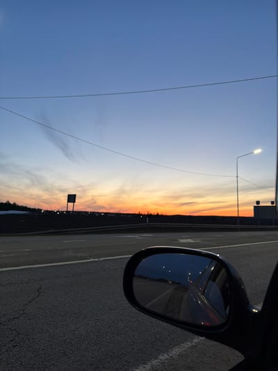 The image is taken from inside a vehicle, potentially a car, while on a bridge or overpass. The right side of the frame features a car's side mirror reflecting a dark highway illuminated by streetlights. To the left, the sky transitions from a light blue at the top to a fiery orange and yellow near the horizon, indicating either a sunrise or sunset. The dark silhouette of trees or a treeline is visible against the horizon. A few utility poles and streetlights punctuate the scene. The image is rotated ninety degrees. Without additional context or geographical landmarks, specifically pinpointing the location within Kuregovo, Russia, is impossible. However, the scene depicts a roadway, likely a highway or main road, possibly on the outskirts of the city, given the presence of a treeline, coupled with an expansive view of the sky and sunset/sunrise.