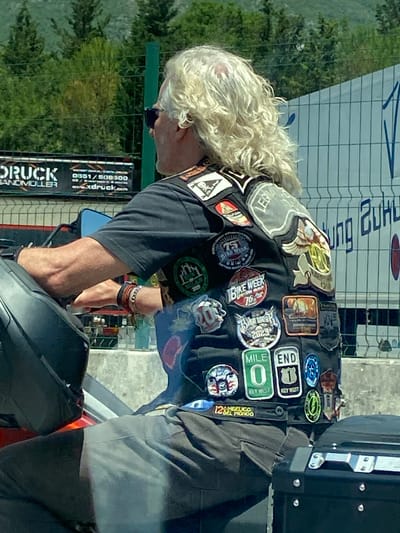 A profile view of a man with long, curly blonde hair and sunglasses is shown from the chest up, riding a motorcycle. He is wearing a dark t-shirt and a black vest adorned with numerous colorful patches. The patches feature various designs and text, including "BIKE WEEK," "MILE 0 KEY WEST," and "END 1 KEY WEST." His left arm is visible, resting on the handlebars, and he is wearing several bracelets on his wrist. The background shows a green fence, trees, and a white structure with blue lettering. The image appears to be taken outdoors on a sunny day. The location is Palje Brdo, Croatia, as indicated by the prompt.