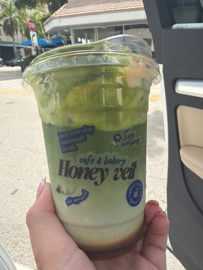 A close-up shot focuses on a clear plastic cup filled with a layered green beverage and topped with a lid. The cup has several stickers on it. One sticker reads "Your everyday handmade break." Below that, a larger sticker says "cafe & bakery Honey veil." Another small sticker depicts a bee and the words "bibi approved." To the right of "Honey veil," a circular sticker with a bee in the center reads "Proudly crafted by ceci & nepi." The cup is held by a hand with manicured nails. In the background, out of focus, is a street scene with palm trees, a stop sign, and buildings. The scene appears to be in South Miami, Florida, given the context of the palm trees and style of buildings. The car door is visible on the right side of the image.