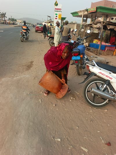 An older woman in a maroon headscarf carries a large, wooden mortar next to motorcycles parked on a dusty roadside. A "POPSY" gas station sign is visible in the background, along with other figures standing nearby. One man is on a moving motorcycle in the background. The scene appears to be in a somewhat rural or less developed area.