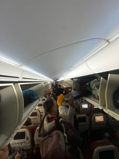The image captures the interior of a commercial aircraft cabin at Paris Charles de Gaulle Airport, located in Tremblay-en-France, France. The scene depicts numerous passengers standing and moving within the central aisle, suggesting boarding or disembarkation. Overhead storage bins line both sides of the cabin; many are open, revealing various pieces of carry-on luggage, including a prominent blue bag and a multi-colored striped bag. Passenger seats with maroon-colored upholstery and individual in-flight entertainment screens are visible on both sides of the aisle, extending towards the rear of the aircraft. In the mid-ground, a man wearing a yellow turban and a light-colored garment stands facing to the right, looking upwards with his right arm raised towards an overhead bin. To his immediate left, another man, seen from behind, with short hair and wearing a dark top over a lighter layer, stands in the aisle, carrying a dark backpack. The cabin is illuminated by artificial overhead lighting, and some personal screens display active content.