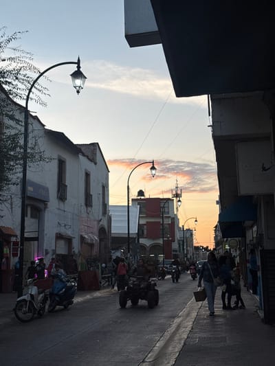 A street scene unfolds with parked motorcycles and ATVs lining the right side. Buildings flank either side, their architecture varying, with some showing signs of age and wear. Streetlights punctuate the scene. Pedestrians stroll further down the street. The sky transitions from a light blue to a soft orange and pink at the horizon.
