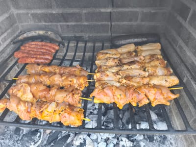 In Campezo/Kanpezu, Spain, a close-up, eye-level shot captures food being grilled on a metal grate over charcoal. The grill is situated within a brick barbecue. On the left side of the grill, there are rows of sausages and some plantains. To the right of the sausages and plantains, there are chicken wings and skewers of marinated chicken pieces. The chicken is seasoned with herbs and spices. The charcoal below is glowing, indicating heat. The scene is brightly lit, likely by natural daylight.