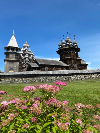 The image shows the Church of the Transfiguration, a wooden church with multiple domes and a bell tower, located in Boyarshchina, Russia. The church is surrounded by a wooden fence and a stone wall. In the foreground, there are pink flowers and green grass. The sky is blue with some clouds. The scene is bathed in bright sunlight, suggesting it is daytime. There are no people or animals visible in the image. The architecture is traditional Russian wooden construction.