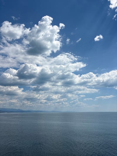The image captures a vast expanse of the sea under a dynamic sky filled with scattered, fluffy white clouds. The horizon is clearly defined, separating the deep blue water from the lighter blue of the sky. In the distance, a faint, mountainous coastline is visible, suggesting a temperate climate. The sea surface is textured with small ripples, indicating a gentle breeze. The overall impression is one of natural beauty and tranquility, with the dramatic cloud formations adding depth and visual interest to the scene. The image does not contain any people, structures, or specific landmarks that would indicate its precise location within Bondarenkovo, Ukraine, beyond the general environmental context of a coastal area.