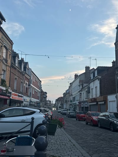 The image depicts a street scene in Noyon, France, likely during the evening. The street is paved with cobblestones and lined with buildings on both sides, featuring a mix of architectural styles. Several cars are parked along the street, including a white hatchback in the foreground, a red hatchback, and a dark-colored SUV. The sky is a pale blue with some wispy clouds and a crescent moon visible. String lights are hung across the street. The overall atmosphere suggests a quiet, possibly closing hour for businesses. Some storefronts are visible with signage in French. A table with chairs and a glass of red liquid is in the lower left corner, suggesting an outdoor seating area.