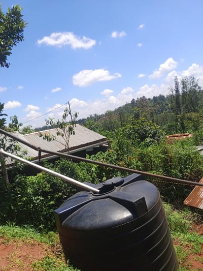 This image depicts an outdoor scene in Kiangochi, Kenya, under a clear blue sky with scattered white cumulus clouds. In the immediate foreground, a black, vertical, ribbed plastic water storage tank occupies the lower right quadrant, with a light-colored pipe extending into its top opening. A darker metal pipe runs horizontally above and behind the tank. The ground slopes downwards from left to right, covered with reddish-brown earth and green grass and weeds. In the mid-ground, dense green vegetation, including bushes and trees, predominates. To the left, a building with a grey corrugated metal roof is partially visible, alongside a small tree with green fruits. Further right, a section of a rusted corrugated metal roof is present. Utility lines are strung across the mid-ground. The background features distant hills and mountains heavily covered in green trees, indicating a forested landscape. No individuals are visible.