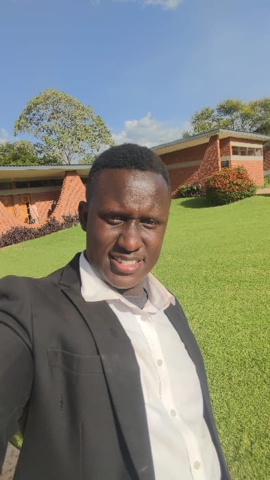 A smiling Black man in a dark suit jacket and white shirt stands outdoors. Behind him, well-maintained green grass covers the ground. A red brick building with small windows is visible, partly obscured by greenery. Above, a tree with light green leaves stands against a blue sky dotted with white clouds. The photo appears to be taken at an angle, slightly tilted to the right.