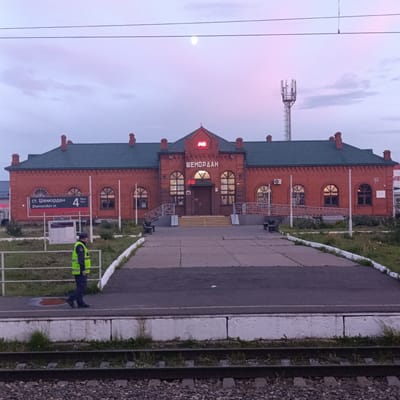 The image depicts a brick train station building in Шемордан, Russia. The building has a dark green roof with several brick chimneys. Above the entrance, the word "ШЕМООРДАН" is visible in red lettering. To the left of the entrance, a sign reads "ст. Шеmордан Shemordan st." with the number "4" and "Путь Track" next to it. A person wearing a high-visibility yellow vest and dark pants is walking on a paved area in front of the station. The scene appears to be at dusk or dawn, with a pale pink and purple sky. There are power lines running across the top of the image and a cellular tower is visible in the distance. Railroad tracks are in the foreground.
