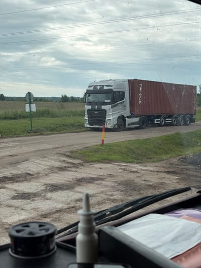 An overcast day is depicted with a silver semi-truck hauling a large, dark red shipping container on a dirt road. The shipping container has "TRITON" printed vertically on its side. The truck is positioned in the middle of the frame, traveling from left to right. A yellow and red striped pole is visible near the truck on the right side of the road. A green signpost with two white rectangular signs is on the left side of the road. The surrounding environment consists of a grassy field and some trees in the background. In the foreground, the interior of a vehicle is visible, including a windshield wiper and dashboard elements. The location is identified as Zarech'e, Russia.