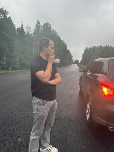 A man stands on a wet, asphalt road in front of the rear of a dark-colored car. The car's taillight is illuminated. The man wears a black t-shirt, light grey jeans, and white sneakers with black stripes. He has his arms crossed and his hand near his mouth, possibly smoking. A forest is visible in the background under a cloudy sky. The location is Михайловка, Russia.
