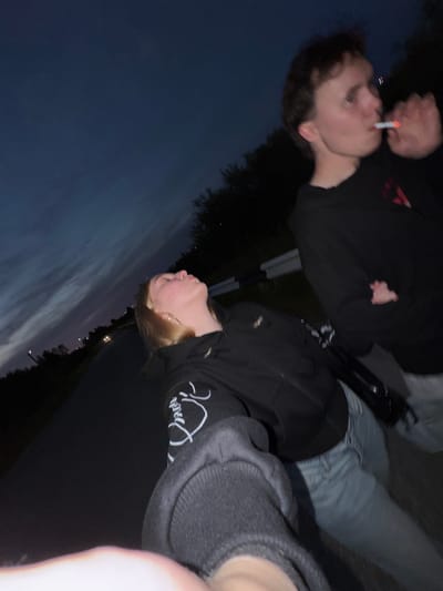 The image shows two individuals standing on a road at dusk, with a dark, cloudy sky overhead. The person on the right is a young male wearing a black hooded sweatshirt and is smoking a cigarette. The person on the left is a young female wearing a black hooded sweatshirt and light blue jeans, looking upwards with her head tilted back. The road appears to be paved and has a guardrail along its side. The background consists of dark trees and distant lights, suggesting an urban or semi-urban environment. The location is identified as Ryabinino, Russia.
