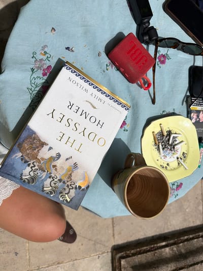 The image captures a still-life scene set in what appears to be an outdoor setting, possibly in Saint-Victor-la-Coste, France. A light blue tablecloth with floral embroidery covers a small table. Various items are scattered on the table, including: a black smartphone, sunglasses, a pack of cigarettes, a small yellow plate filled with cigarette butts, a mug, a red JBL speaker, and a black DJI Osmo Pocket camera. Central to the composition is a book, "The Odyssey" by Homer, translated by Emily Wilson. A person, presumably seated, is partially visible in the bottom right corner, with a glimpse of their leg and the edge of a white lace garment. The environment appears to be a sunlit outdoor patio or terrace with the edge of a stone structure visible in the upper right corner.