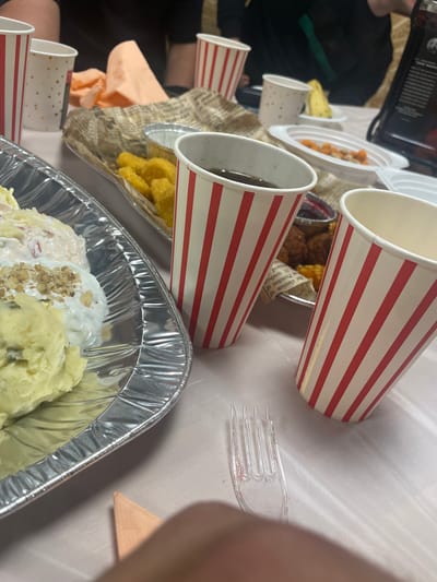 The image depicts a dining setting with various food items and disposable tableware. There are paper cups with red and white stripes, some containing liquid. Food is served in aluminum foil containers, including what appears to be mashed potatoes with toppings. There's also what looks like fried food in another container. A plastic fork is visible. The setting is a table covered with a white tablecloth. The image does not provide information that connects it to Mirkovo, Bulgaria. There is no presence of people or subjects identified by name, action or interaction. The environment shown is a table with food.