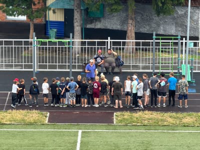 A group of children, possibly a sports team, stands on a running track facing away from the camera. They are gathered behind a white fence with green accents. Two adults, one in a red cap and a black t-shirt, the other in a gray t-shirt and dark pants, are seated on a bench to the right of the fence. A third adult in a blue t-shirt and light pants stands to the left of the fence, facing the children. The background features a building with a brick and corrugated metal exterior, along with trees. A green trash bin is visible near the building. The scene appears to be an outdoor sports facility. The image is likely taken in Tashtagol, Russia, given the context.