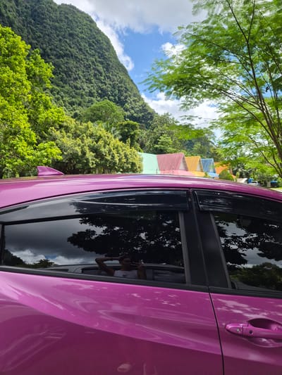 A vibrant pink car is parked in a scenic location, likely a resort or roadside stop, in Phang-nga, Thailand. The image is taken from a low angle, looking up towards a lush green mountainside covered in dense foliage. The sky is a mix of bright blue with scattered white clouds, suggesting it is daytime with good weather.

In the foreground, the side of the pink car dominates the right half of the frame. Its glossy surface reflects the surrounding environment, including trees and the sky. A black window trim and a silver door handle are visible.

To the left of the car, a line of colorful buildings with pitched roofs can be seen nestled against the base of the mountain. These buildings, painted in shades of yellow, orange, red, green, and blue, suggest a tropical resort or a collection of chalets.

Further up the mountainside, thick green vegetation covers the slopes, indicative of a tropical climate. A large tree with feathery green leaves hangs over the scene from the top left corner.

Reflected in the car window, a person can be seen leaning out of the car, holding up a hand, possibly taking a selfie or waving. Their posture and the slight smile visible in the reflection suggest a relaxed and happy mood, enjoying the beautiful surroundings. The overall impression is one of a leisurely vacation in a picturesque tropical landscape.