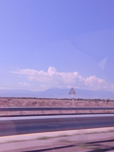 This is a landscape photograph taken from a moving vehicle, likely on a highway. The sky is a pale blue with some wispy clouds, and the horizon is dominated by a range of distant, hazy mountains. In the foreground, a multi-lane highway is visible, with guardrails and clear lane markings. The landscape flanking the highway appears arid and sparsely vegetated, with low hills and some scattered trees. A blurred traffic sign is visible on the left side of the road. The overall impression is one of a vast, open, and somewhat desolate environment. The location in the city of Nurly, Kazakhstan, is not explicitly indicated by any visual cues within the image itself.