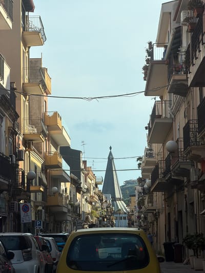The image depicts a narrow street in Chianchitta, Italy, densely lined with multi-story buildings. The perspective is tilted upwards, emphasizing the verticality of the architecture. The buildings are a mix of pale yellow, beige, and light brown hues, featuring balconies, air conditioning units, and various architectural protrusions. The sky is visible between the buildings, with a tall spire or tower-like structure partially visible in the distance. The street is congested with parked and moving vehicles, including a yellow car prominent in the foreground. A blue and white parking sign is visible on one of the buildings. There is a sense of urban density and the composition suggests a cramped, bustling environment. No people are clearly visible in the scene.