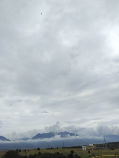 A wide-angle, eye-level shot captures a hazy, overcast sky over a rural landscape. In the upper portion of the frame, a single, dark bird is visible in flight against the diffused light of the clouds. The middle ground features a range of mountains partially obscured by wispy clouds that cling to their slopes. The foreground shows a flat, grassy plain with scattered trees. To the right of the frame, an industrial complex with several buildings, transmission towers, and power lines is situated near the base of the mountains. The overall impression is one of a quiet, possibly industrial, rural setting under a muted sky.
